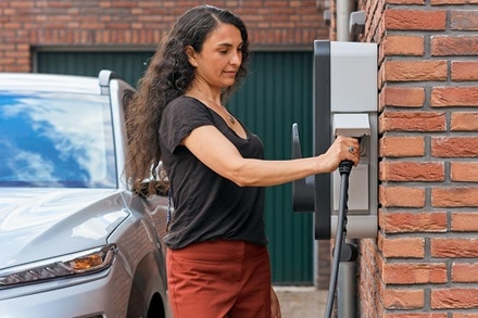 Femme qui branche son câble sur une borne de recharge witty start de Hager montée sur le mur en briques rouges d'une maison