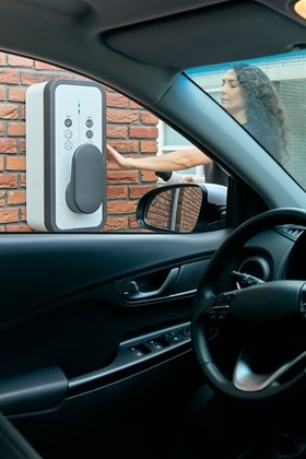 Femme rechargeant sa voiture sur une borne de recharge witty start de Hager fixée sur un mur en brique vue de l'intérieur d'un véhicule électrique, fenêtre conducteur ouverte