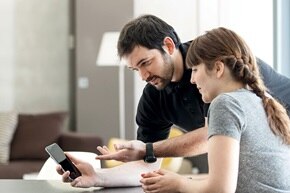Couple qui se renseigne sur le Diagnostic Electrique Obligatoire sur leur smartphone dans leur salon. Diagnostic électrique, DEO, mise aux normes, normes électriques, solutions Hager, solutions connectées