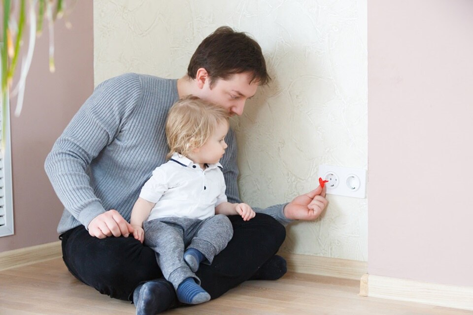 Papa, avec son bébé dans les bras, qui installe un cache de protection dans les prises électriques