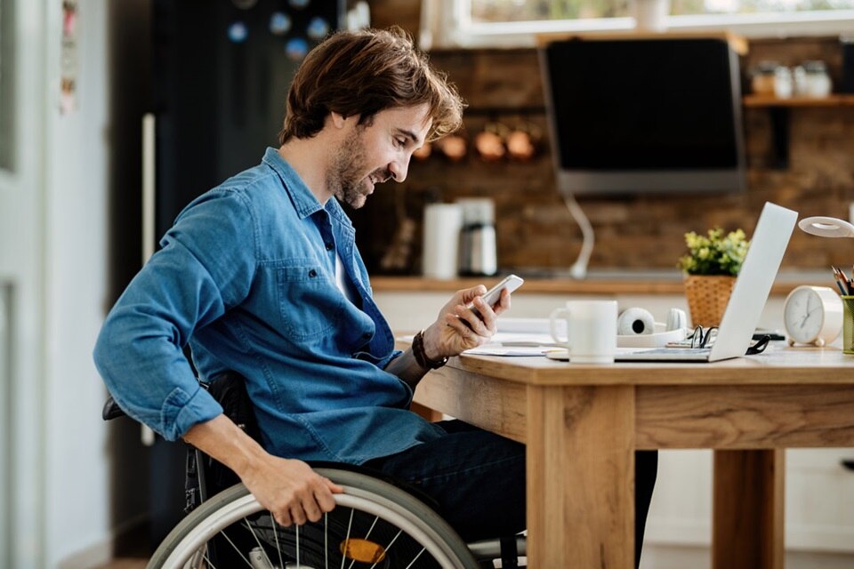 homme souriant, en télétravail dans sa cuisine, sur un fauteuil roulant qui consulte son smartphone