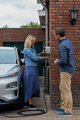 Couple rechargeant leur véhicule électrique à l'aide d'une borne de recharge witty de Hager fixée sur le mur en briques rouges de la maison