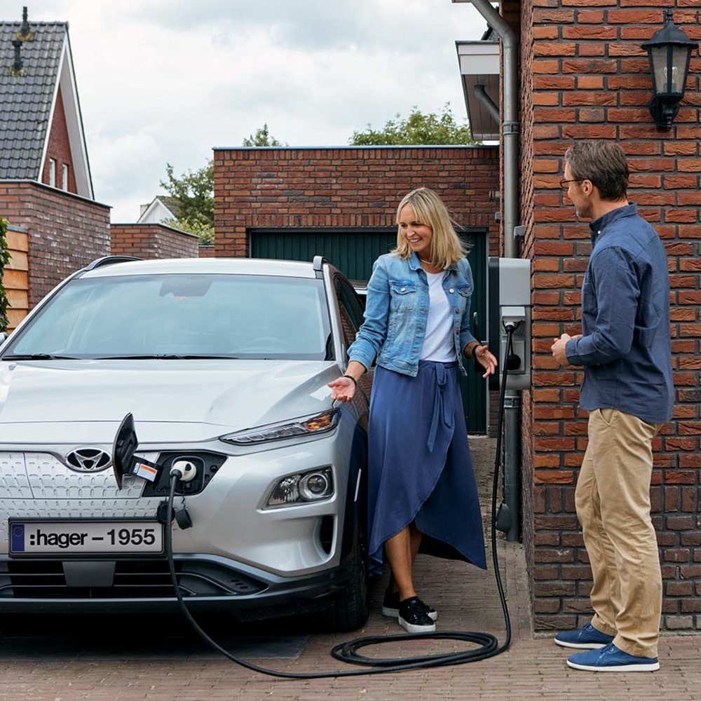 Couple rechargeant leur véhicule électrique à l'aide d'une borne de recharge witty de Hager fixée sur le mur en briques rouges de la maison