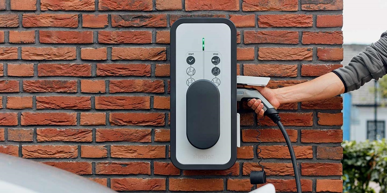 Homme qui recharge son véhicule électrique à l'aide d'une borne de recharge witty de Hager fixée sur le mur en briques rouges d'une maison