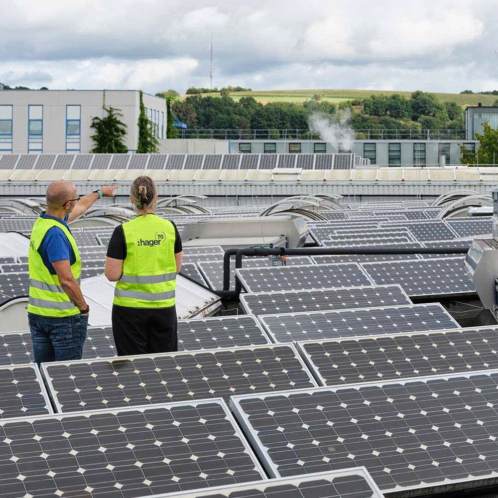 Two people on rooftop with solar panels, buildings and wind turbines in background