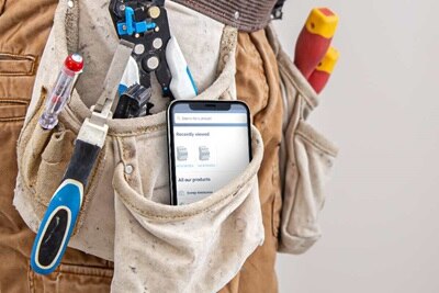Close-up of a professional's tool belt with electrical tools and a smartphone with an electrical distribution product page on the screen
