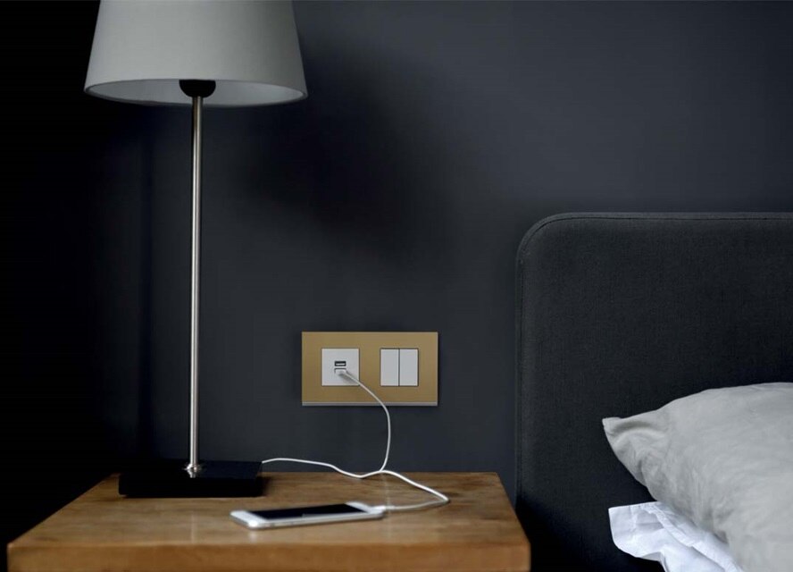 Chambre moderne avec une lampe sur une table de chevet, un smartphone en charge, et un interrupteur lumineux et une prise murale