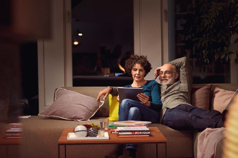 Deux personnes se détendant dans un salon la nuit, l'une utilisant une tablette, éclairage ambiant provenant de luminaires modernes
