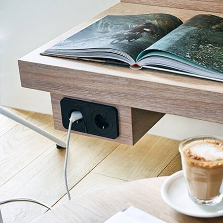 Prise électrique intégrée dans une table en bois avec un cordon branché, à côté d'un magazine ouvert et d'une tasse de café
