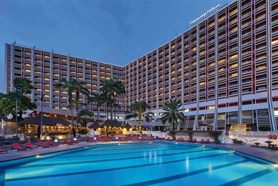 Extérieur de l'hôtel Transcorp Hilton au crépuscule avec fenêtres illuminées, zone au bord de la piscine et palmiers