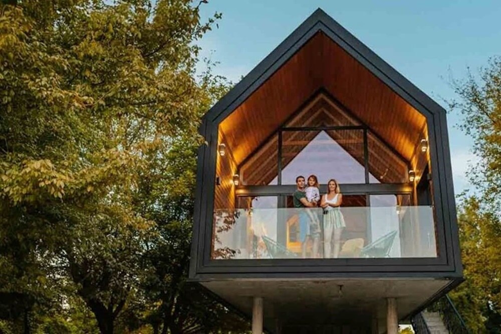 Casa costruita con materiali sostenibili in un contesto eco-compatibile. Affacciato al balcone una famiglia composta da padre, madre e figlio.