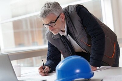 Ingegnere progettista o architetto che esamina piani di costruzione su una scrivania in un ufficio luminoso, con un elmetto di sicurezza blu in primo piano.