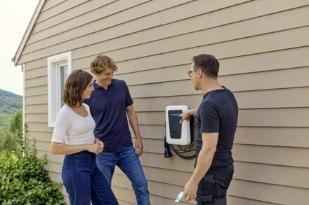 Installatore esperto mostra a una coppia come usare la colonnina di ricarica witty one Hager a parete, con frontalino bianco, installata in ambito residenziale.