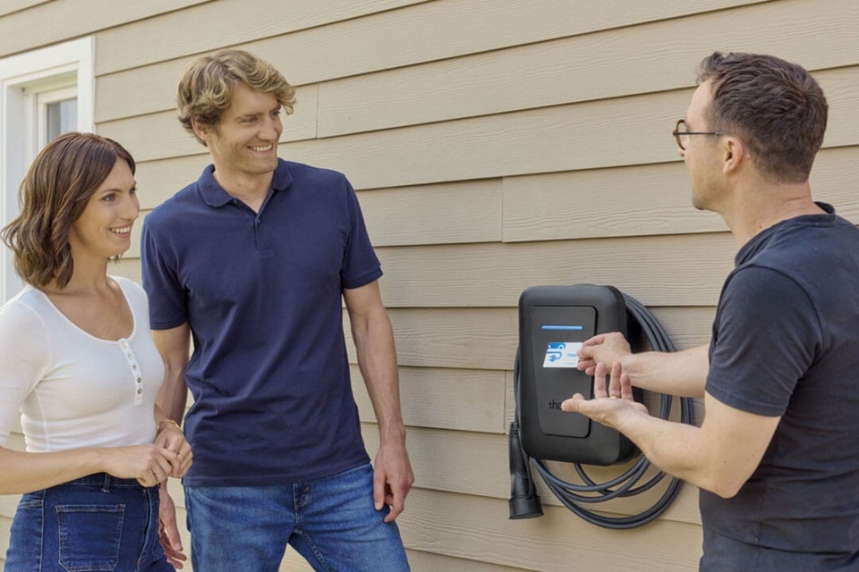 Installatore spiega il funzionamento della colonnina di ricarica per veicoli elettrici witty one Hager a una coppia di clienti, davanti a un'abitazione. La colonnina è installata a parete, con cavo integrato e badge RFID per l'attivazione, evidenziando facilità d'uso e design compatto.