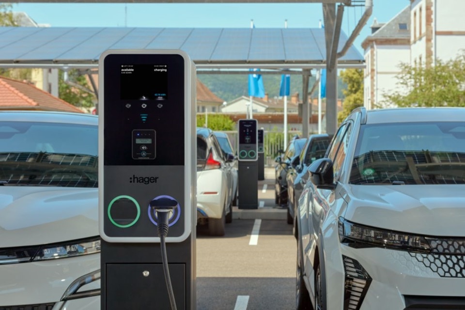 Parcheggio parzialmente coperto per auto elettriche con pannelli solari sul tetto. In primo piano due auto, al centro una colonnina di ricarica witty Park 2 con due punti di ricarica, uno con cavo collegato. L’anello luminoso attorno al connettore più vicino è blu, indicando lo stato di ricarica