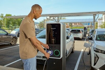 Parcheggio parzialmente coperto per auto elettriche con pannelli solari sul tetto. In primo piano un uomo intento a collegare la presa sulla colonnina di ricarica witty park 2. L’anello luminoso attorno al connettore più vicino è blu, indicando lo stato di ricarica