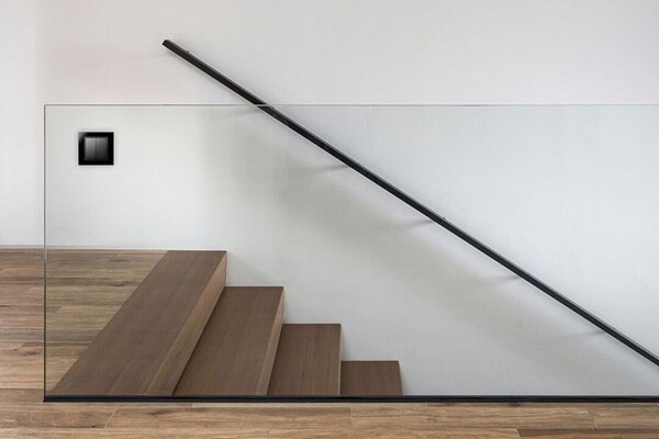 Esempio di installazione del pulsante doppio Berker K.1 grigio con cornice nera su un pannello di vetro trasparente accanto alle scale in legno nel corridoio