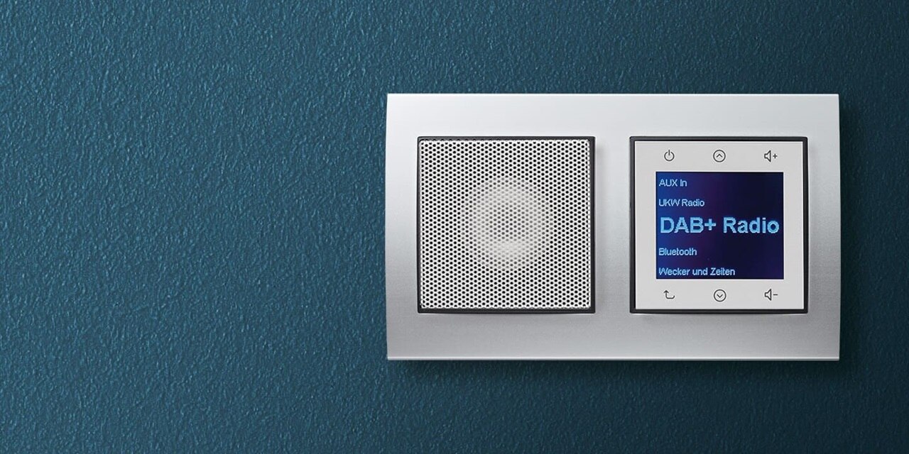 Esempio di installazione di una radio Berker da incasso con display, comandi e altoparlante in bianco con cornice in alluminio su una parete blu