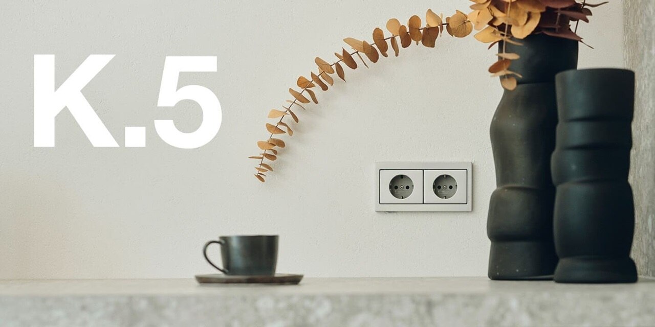  Vista frontale di una stanza con complementi d'arredo neri con sulla parete con doppia presa Berker K.5 in argento, accanto la scritta K.5
