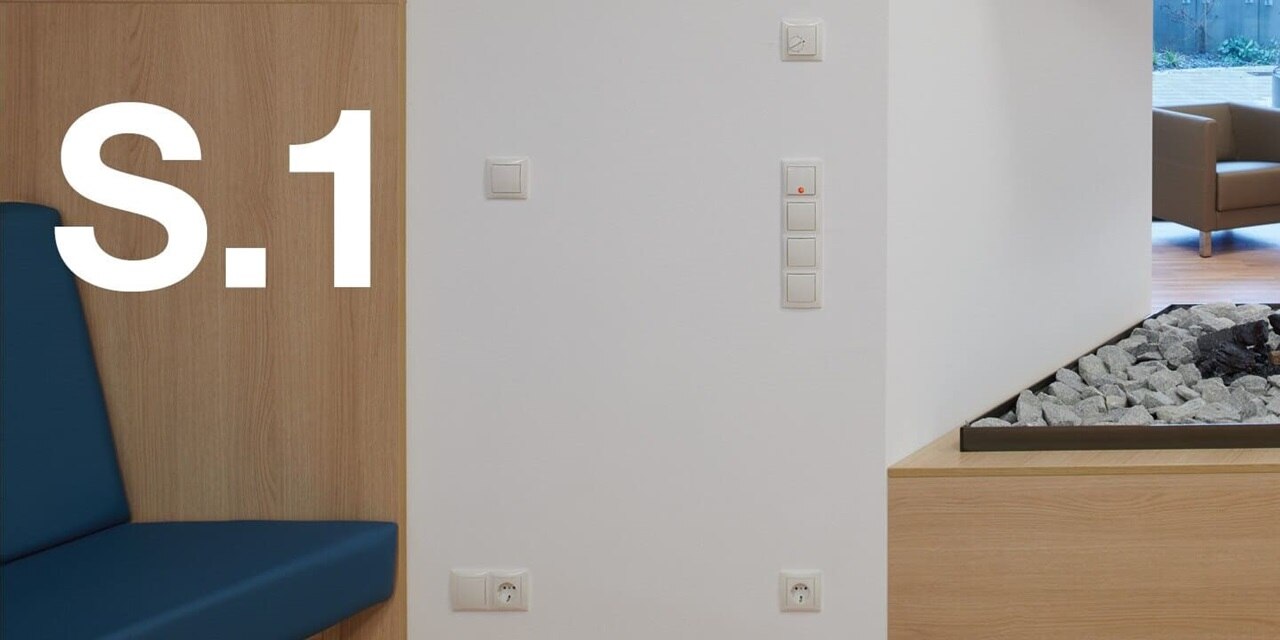  Vista di una parete di un soggiorno con poltrone blu e arredi in legno. Sulla parete sono visibili interruttori e prese installatidella gamma di interruttori Berker S.1 di colore bianco, con la scritta S.1 in evidenza a sinistra