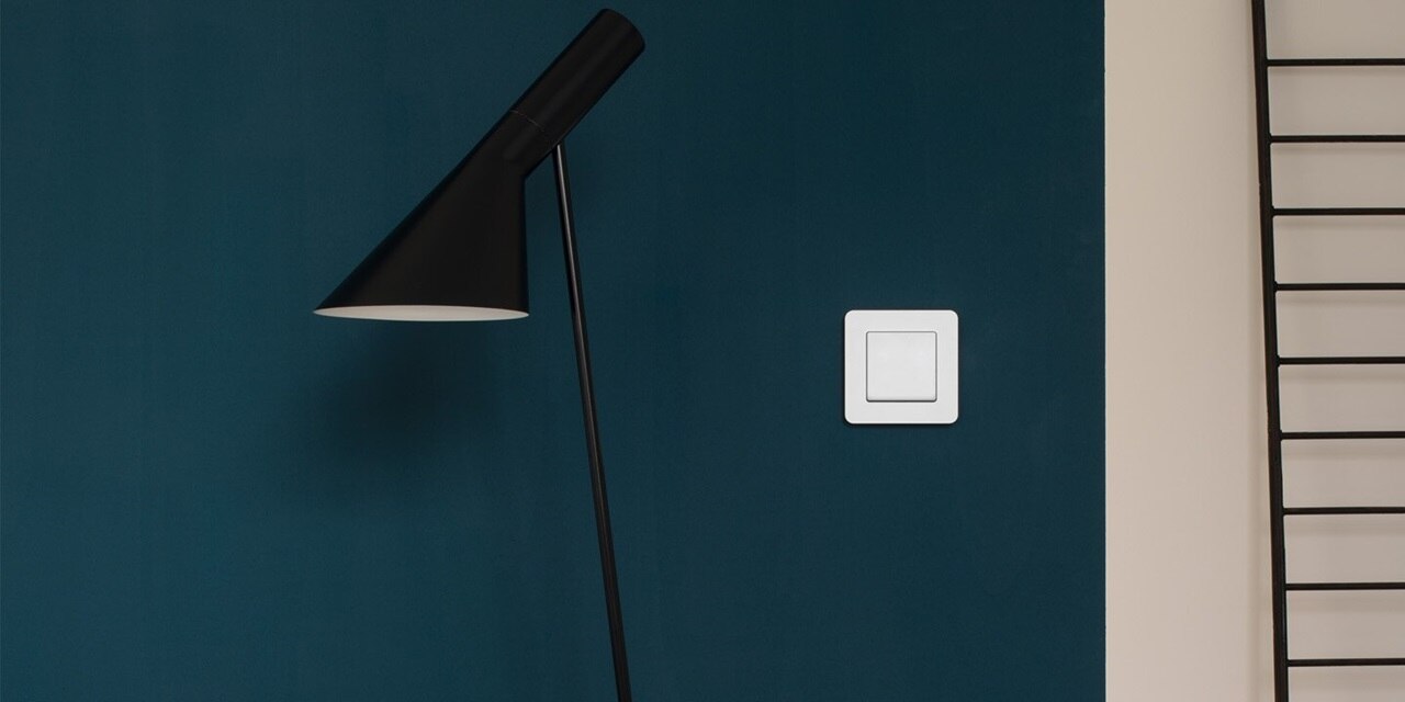 Esempio di installazione di un interruttore della luce Berker Q.7 con bilanciere singolo e cornice bianca su una parete blu accanto a un divano.