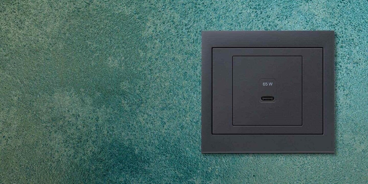 Vista frontale della presa di ricarica USB Berker K.1 da 65 watt con una porta di ricarica USB-C e cornice antracite su sfondo screziato blu-verde.