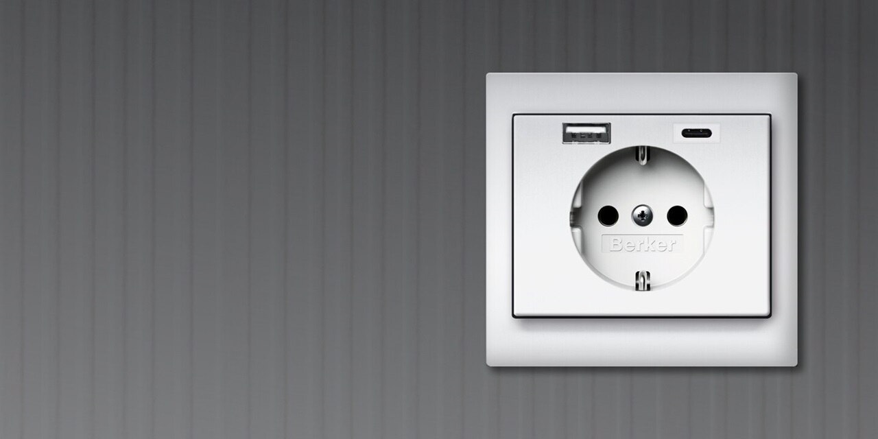 Vista frontale della presa tipo shuko Berker K.1 con due porte di ricarica USB, una USB-A e una USB-C, con cornice bianca su sfondo grigio