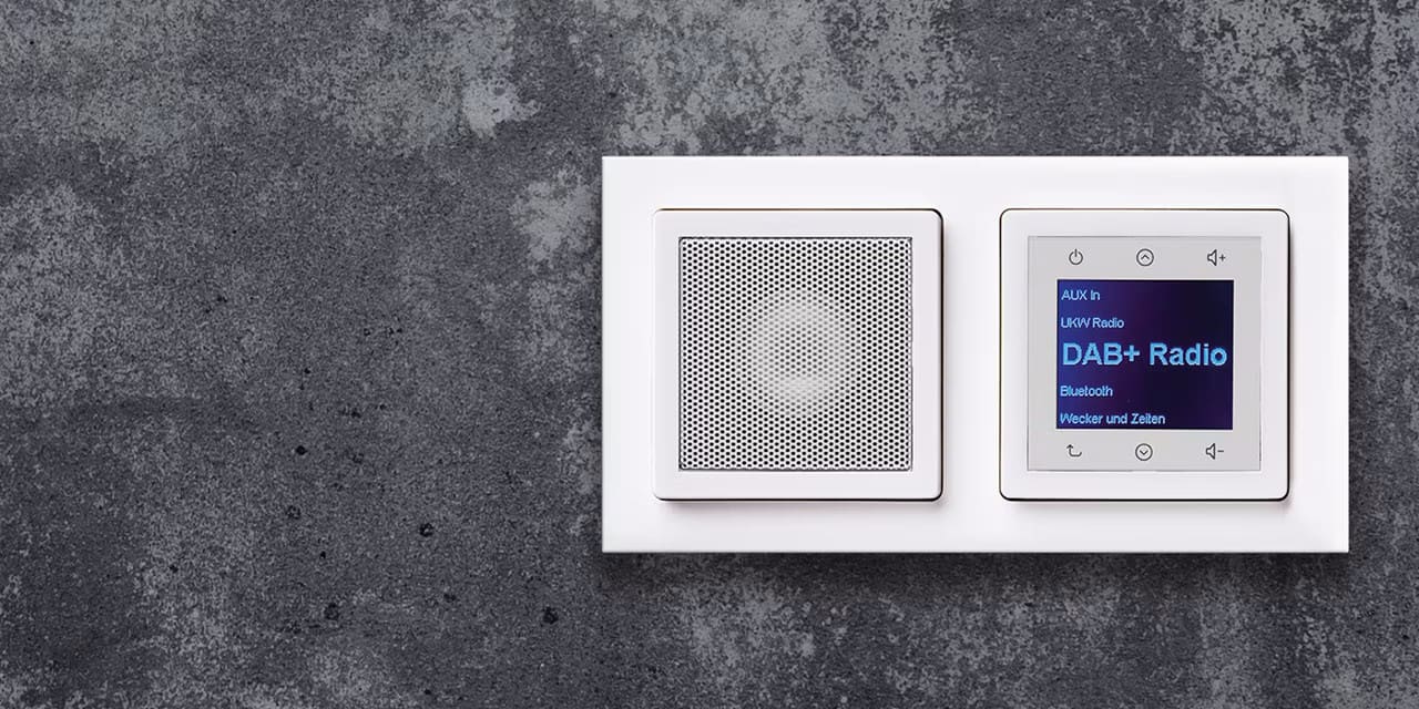 Esempio di installazione della radio Berker da incasso con display, elementi di comando e altoparlante con cornice in alluminio della serie di interruttori e prese Q.3 su una parete con intonaco fine grigio