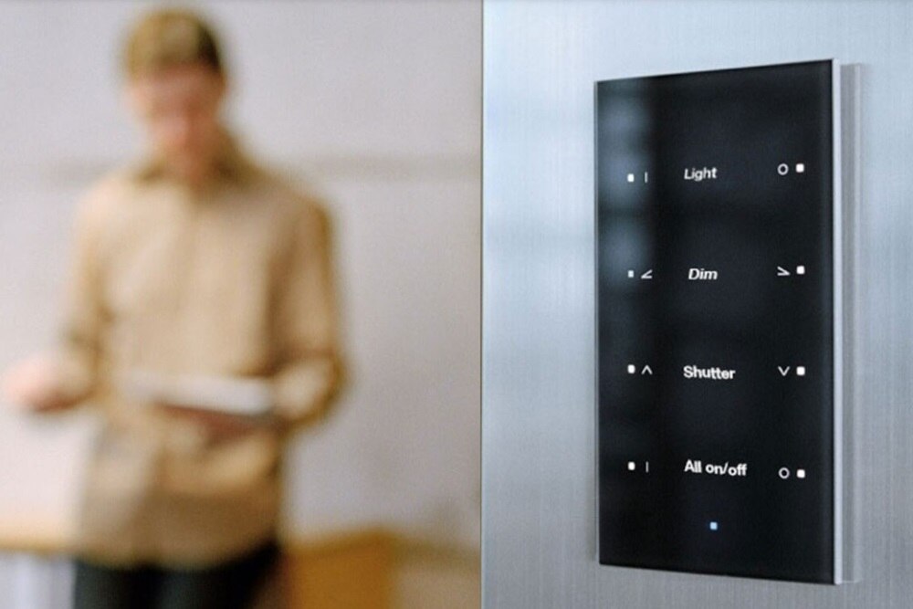 Primo piano di un sensore vetro KNX montato a parete con evidenziate etichette personalizzate e associate ad ogni pulsante. Sullo sfondo, sfuocata è visibile una presenza maschile.