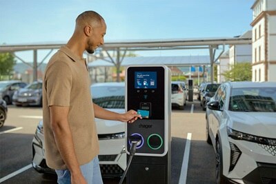 Parcheggio parzialmente coperto per auto elettriche con pannelli solari sul tetto. In primo piano un uomo intento a passare la carta RFID sulla colonnina di ricarica witty Park 2. L’anello luminoso attorno al connettore più vicino è blu, indicando lo stato di ricarica