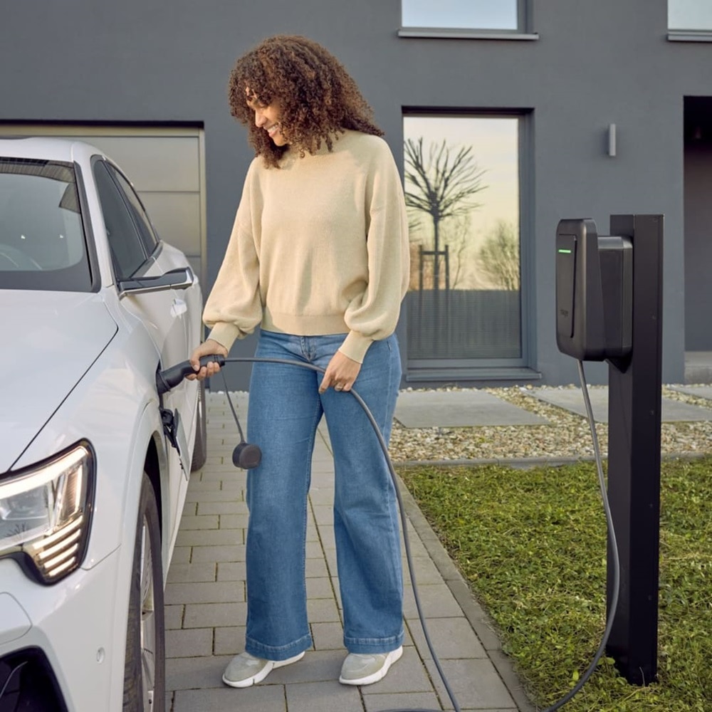 Donna sorridente in piedi intenta a ricaricare il veicolo elettrico con l’energia generata dalla colonnina di ricarica witty plus alle sue spalle, installata in un parcheggio aziendale