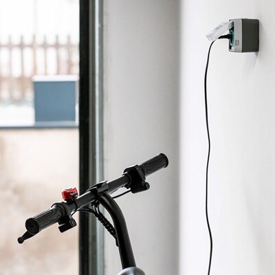 Monopattino elettrico collegato a una presa di ricarica witty prise montata a parete in un garage domestico. L'immagine mostra il cavo inserito nel mezzo e la presa installata su una parete bianca.
