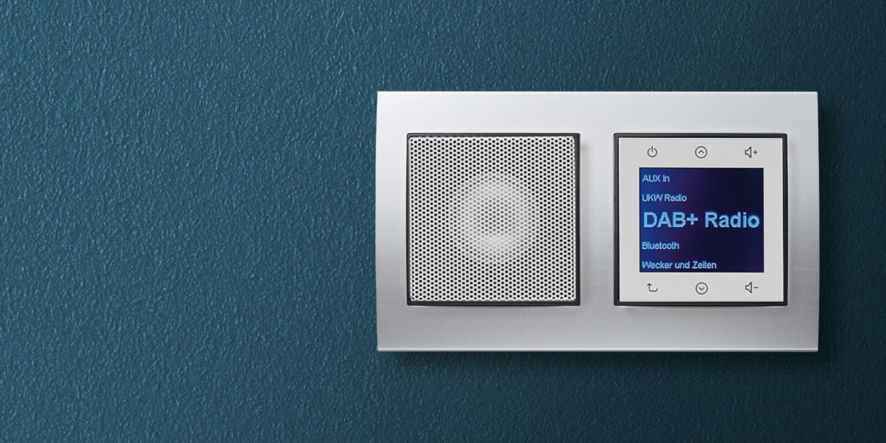 Esempio di installazione di una radio Berker da incasso con display, comandi e altoparlante in bianco con cornice in alluminio su una parete blu