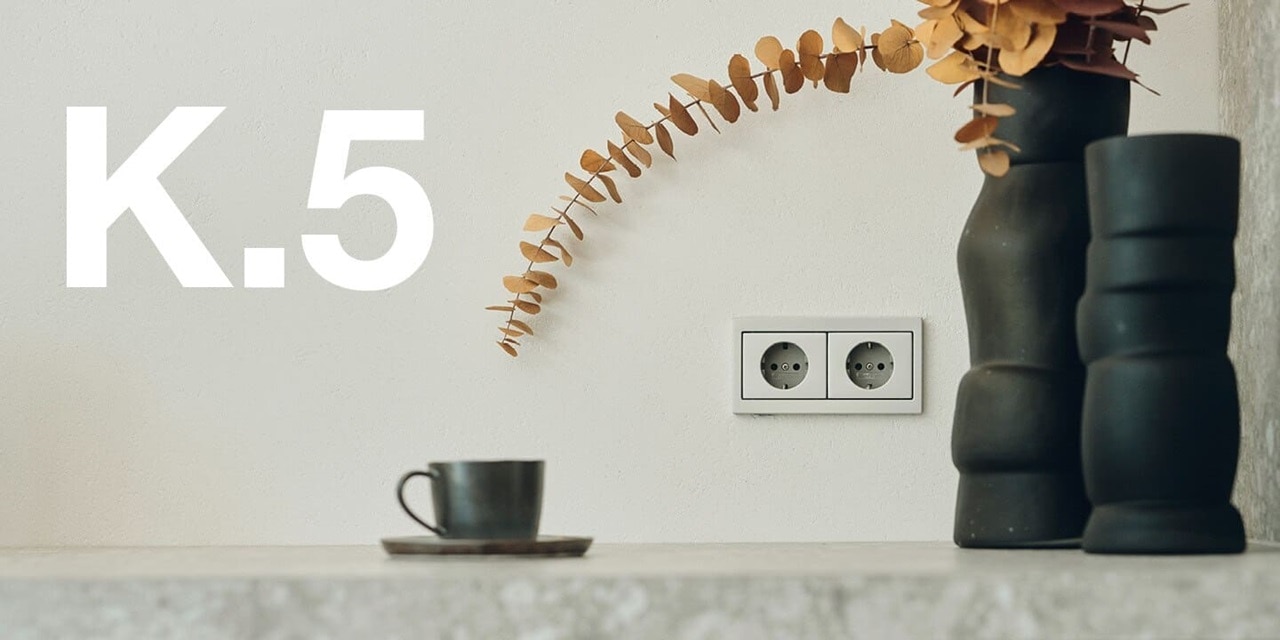 Vista frontale di una stanza con complementi d'arredo neri con sulla parete con doppia presa Berker K.5 in argento, accanto la scritta K.5