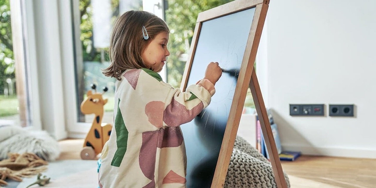 Bambina che disegna su una lavagna con prese e interruttori Berker K.1 sullo sfondo