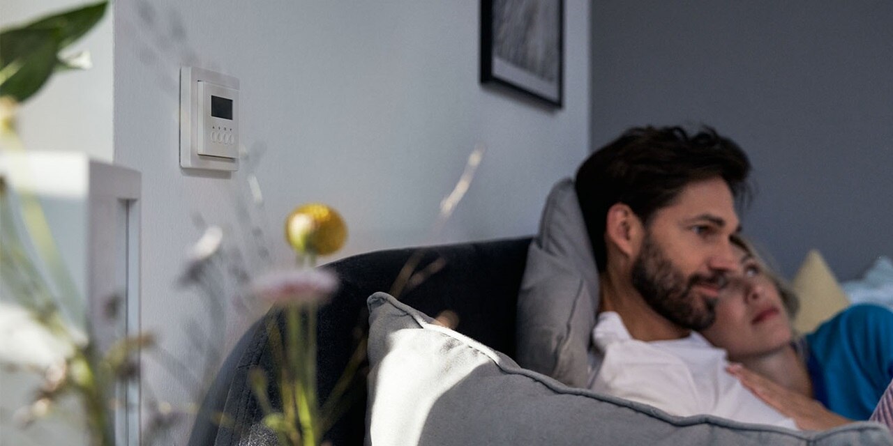 Coppia che si rilassa sul divano con un termostato Berker Q.1 a parete sullo sfondo