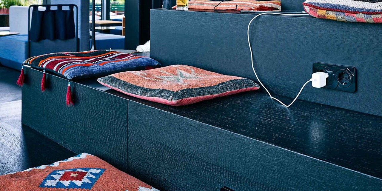 Esempio di installazione di prese Berker R.1 con elemento centrale rotondo e cornice arrotondata in nero su legno color denim della zona salotto dell'hotel