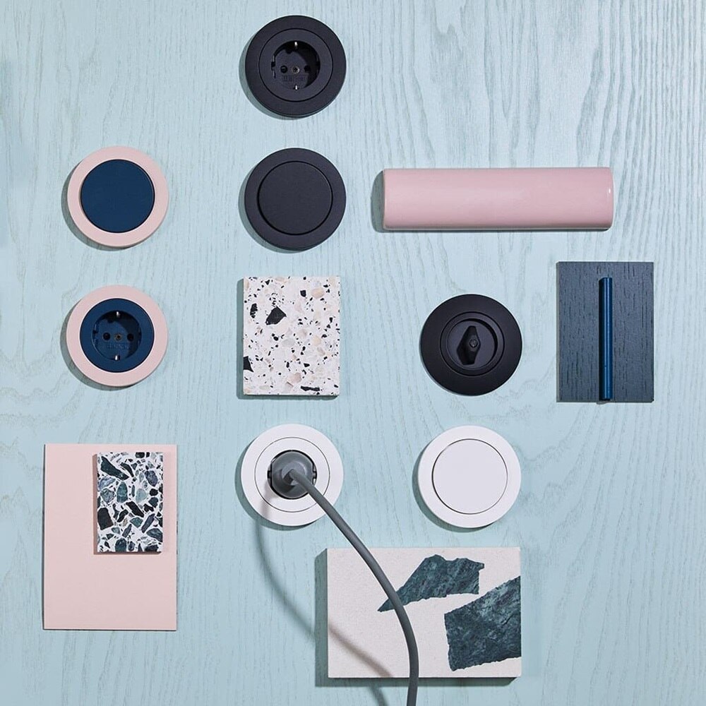 Serie limitata di prese e interruttori Berker Manufaktur nelle varianti rosa e verde, esposti su un tavolo in legno verde acqua insieme a campioni e elementi d’arredo.