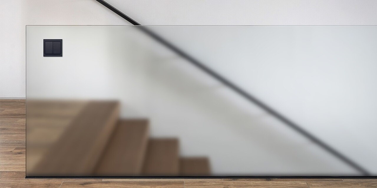 Esempio di installazione del pulsante doppio Berker K.1 con cornice in antracite su pannello in vetro satinato accanto alla scala in legno nel corridoio