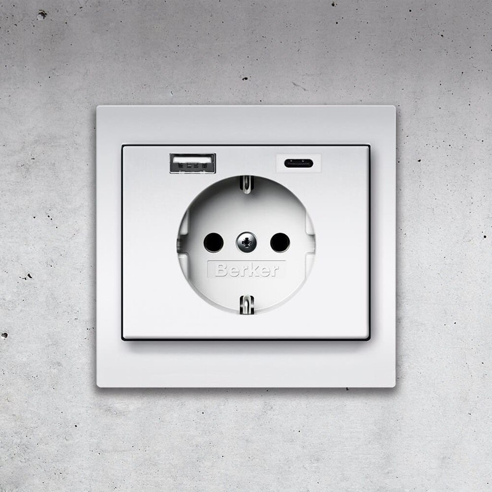 Vista frontale della presa tipo shuko Berker K.5 con 2 porte di ricarica USB - 1x USB-A e 1x USB-C - e cornice in alluminio su un muro di cemento