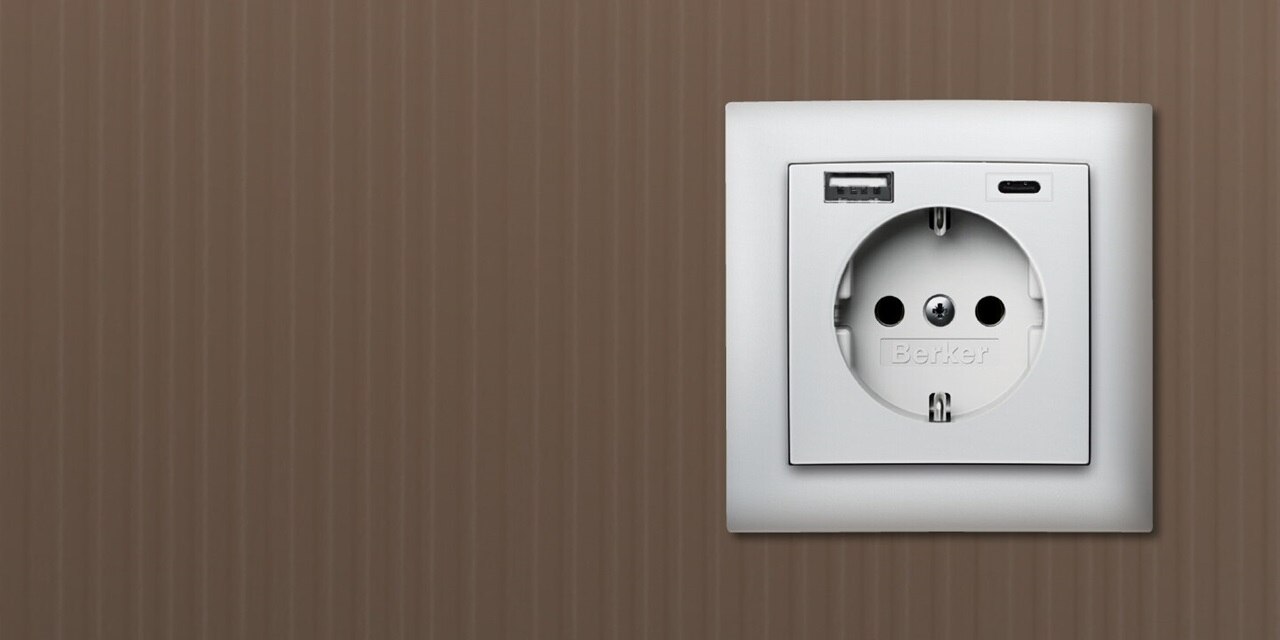 Vista frontale della presa tipo shuko Berker S.1 con due porte di ricarica USB, una USB-A e una USB-C, con cornice bianca su sfondo marrone