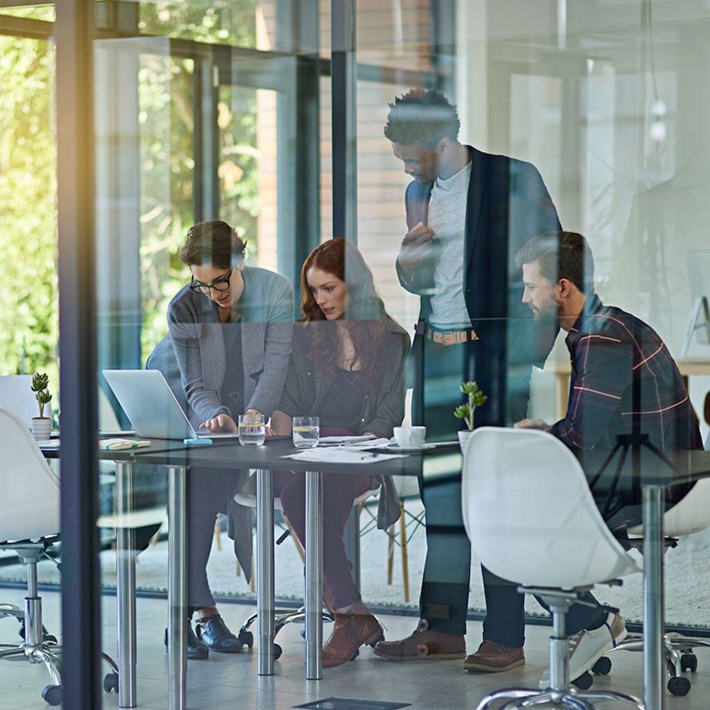 Due donne sedute ad una scrivania davanti ad un notebook e un uomo in piedi dietro di loro, all’interno di una sala riunioni chiusa da una parete in vetro 