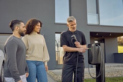 Drie mensen bij een moderne elektrische laadpaal: één persoon toont hoe het werkt, terwijl de anderen geïnteresseerd toekijken. Op de achtergrond een modern gebouw
