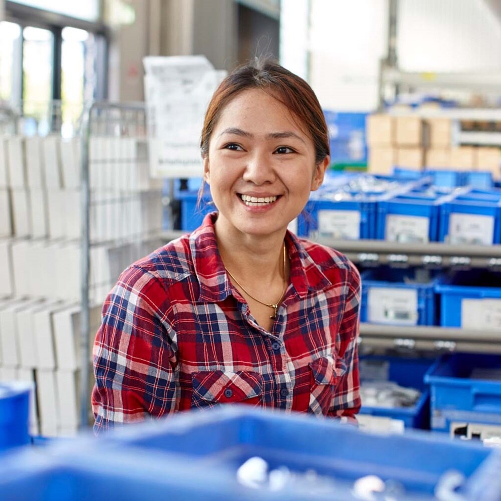 Persoon in geruite blouse staat in een opslagruimte met rekken vol blauwe bakken en stapels witte dozen, passend binnen een logistieke werkomgeving.