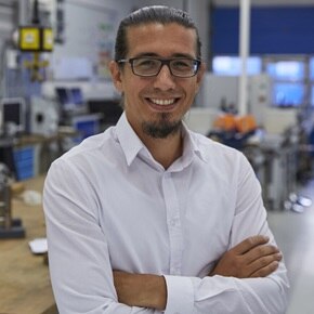 Persoon met gekruiste armen in een technische werkplaats met computers en machines op de achtergrond.