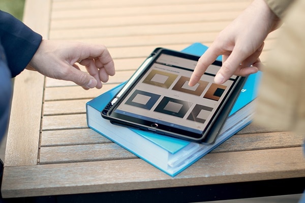 Twee handen bedienen een tablet met kleurstalen op het scherm, liggend op een blauw boek op een houten tafel