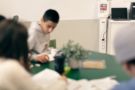 Groep mensen studeert of werkt samen rond een tafel in een klaslokaal, met boeken, papieren en een whiteboard op de achtergrond.