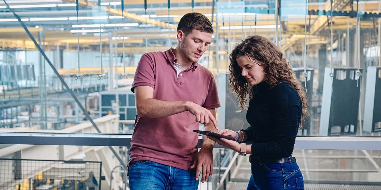 Twee personen in een moderne industriële omgeving bekijken samen informatie op een tablet; één wijst naar het scherm terwijl de ander het apparaat vasthoudt. Op de achtergrond zijn machines en werkplekinfrastructuur zichtbaar.