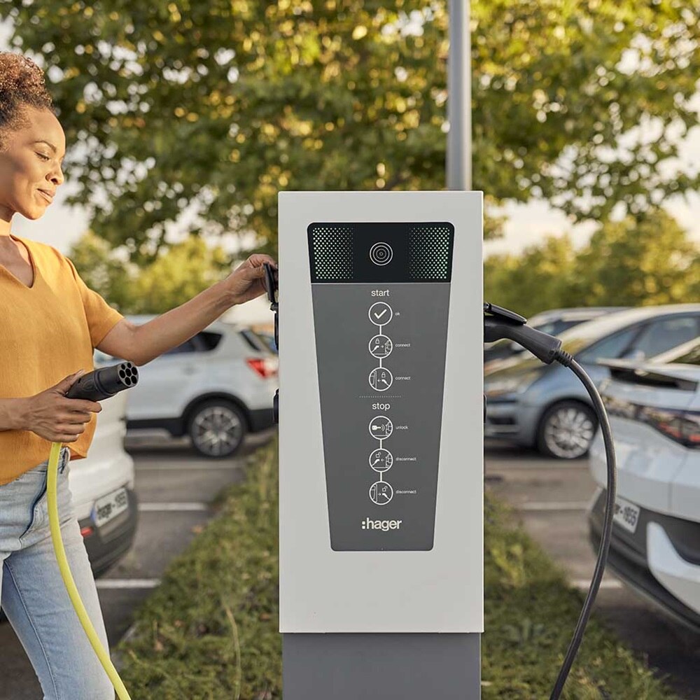 Persoon bedient een elektrische laadpaal met zichtbaar bedieningsscherm en merk “hager”; auto’s op de achtergrond duiden op een publieke laadzone.