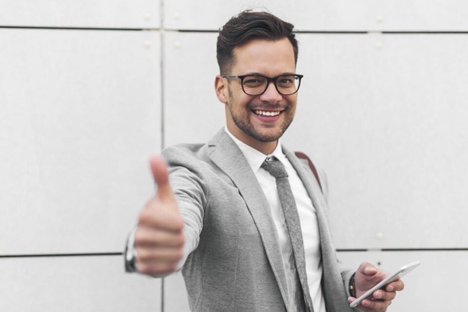 Uśmiechnięta osoba w biznesowym stroju pokazująca kciuka do góry trzymając smartfona